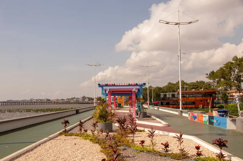 Paseo del Píer do Santa Inês con pérgola colorida, jardín de grava y plantas, postes de luz y un restaurante flotante de madera al fondo.