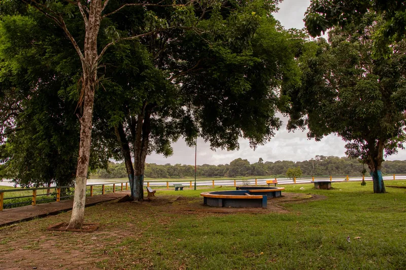 Praça arborizada da Orla de Pracuúba com bancos e canteiros circulares, guarda-corpo amarelo e o rio ao fundo.