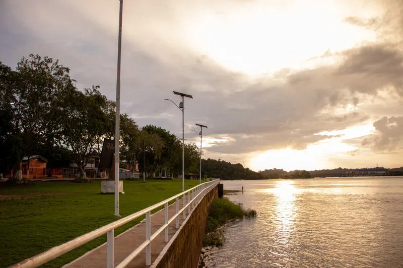 Calçadão com guarda-corpo branco na orla de Ferreira Gomes, gramado e postes, com o sol se pondo sobre o rio