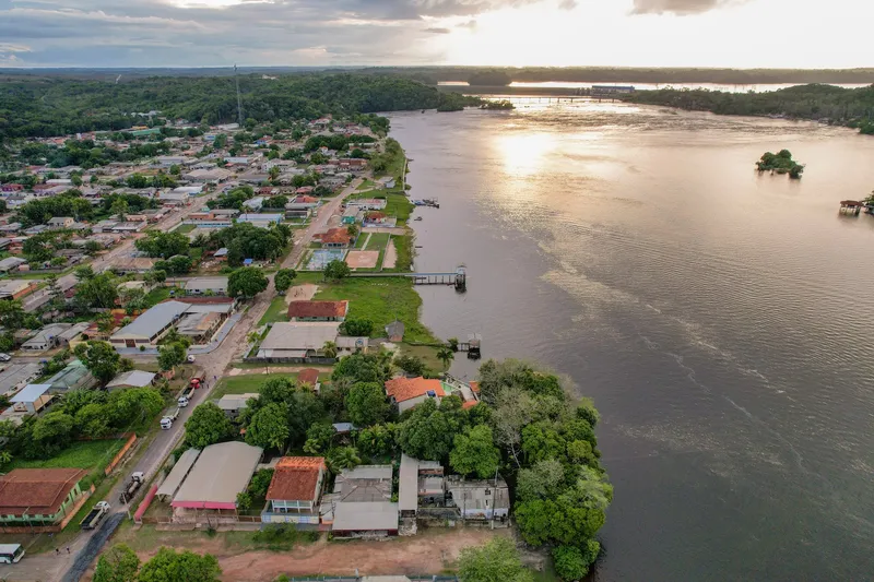 Vista aérea da orla de Ferreira Gomes ao entardecer: casas à margem do rio largo, trapiches e mata ao fundo